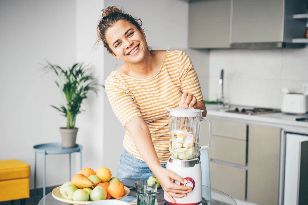 Author Ava Sinclair smiling at the camera while adding fresh fruit to a blender for an easy weeknight recipe.