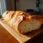 A freshly baked loaf of homemade french bread with a golden, crackly crust resting on a wooden cutting board.