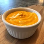 A close-up of creamy, vibrant orange homemade nacho cheese sauce served in a small white fluted ramekin.