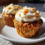 Close-up of two delicious carrot cake muffins topped with thick cream cheese frosting and shredded carrot garnish.