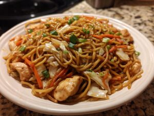 A close-up of a plate piled high with homemade chicken chow mein recipe, featuring noodles, chicken pieces, carrots, and green onions.