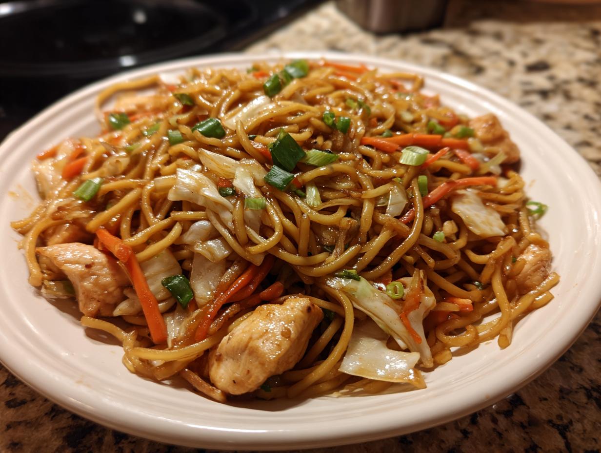 A close-up of a plate piled high with homemade chicken chow mein recipe, featuring noodles, chicken pieces, carrots, and green onions.