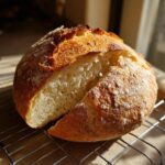 A freshly baked, round loaf of Italian bread with a golden, cracked crust, resting on a cooling rack with one slice cut away.