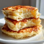 A close-up of three incredibly fluffy pancakes stacked, showing their airy interior and golden-brown edges, drizzled with syrup.