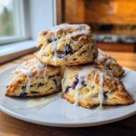 A stack of three freshly baked blueberry scones drizzled generously with white lemon glaze, sitting on a white plate.