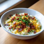 A close-up of a white bowl filled with creamy Loaded Baked Potato Soup, topped generously with shredded cheddar cheese, bacon bits, and fresh chives.