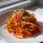 A bright mound of raw carrot salad made from shaved carrots, topped with black and white sesame seeds and green onions.