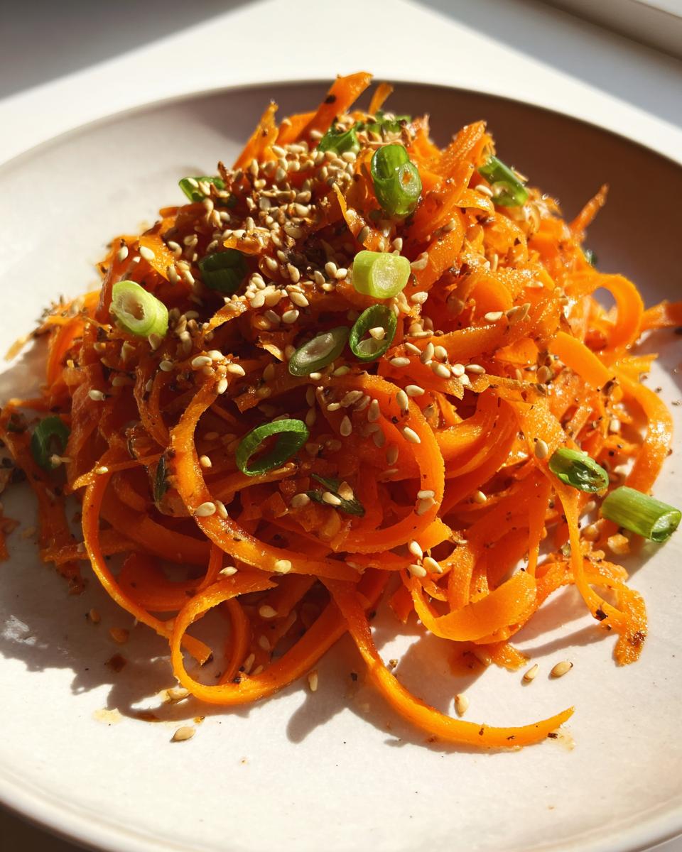 A close-up of a vibrant raw carrot salad made with spiralized carrots, topped with toasted sesame seeds and sliced green onions.