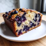 A thick slice of moist blueberry banana bread, studded with fresh blueberries, served on a white plate.