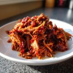 A close-up of juicy, saucy Slow Cooker Pulled Pork shreds piled high on a white plate.