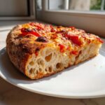 Close-up of a thick slice of pizza made with sourdough discard pizza dough, showing a highly aerated, open crumb structure.