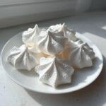 A cluster of perfectly piped, star-shaped white meringue cookies resting on a small white plate near a window.
