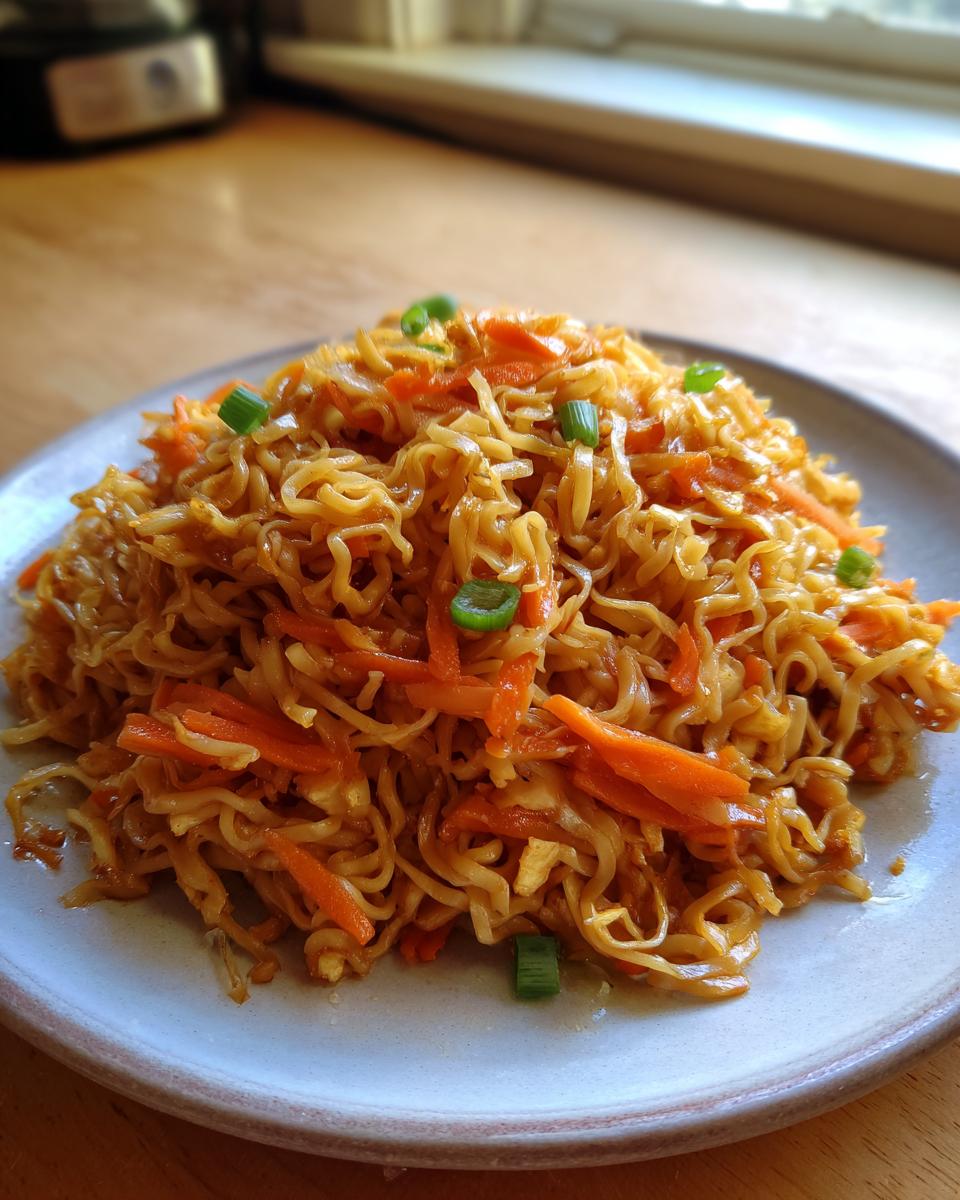 A plate piled high with glossy, savory fried noodles mixed with shredded carrots and topped with green onions.
