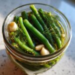 Close-up overhead view of bright green dilly beans packed in a glass jar with garlic cloves, fresh dill, and pickling spices.