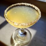A bright yellow lemon drop cocktail in a coupe glass with a coarse sugar and citrus zest rim, catching sunlight.