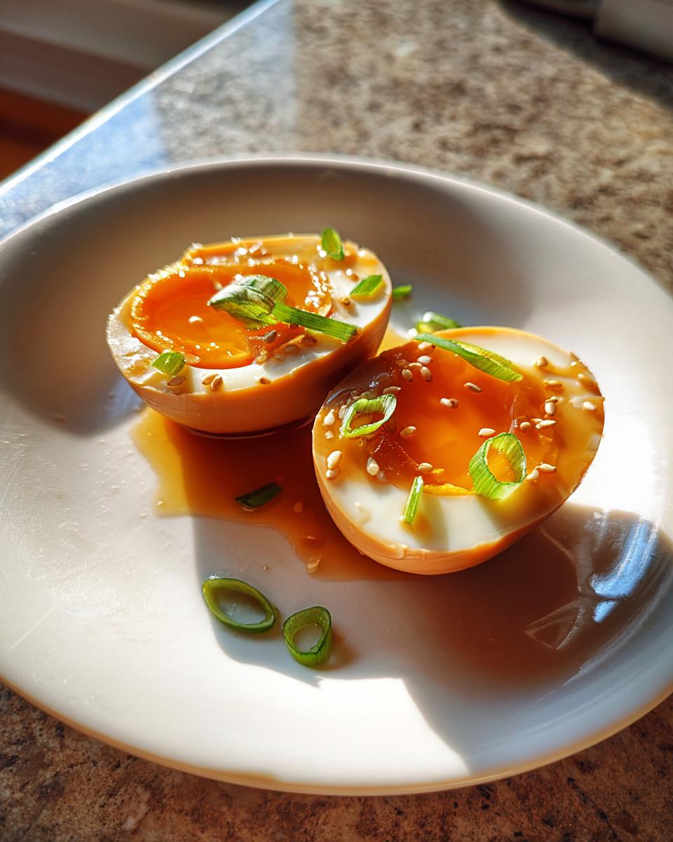 Two halves of a marinated eggs with jammy orange yolks, drizzled with sauce and topped with sesame seeds and scallions.