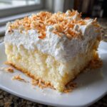 A moist slice of coconut poke cake topped with thick whipped cream and toasted coconut flakes.