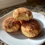 Close-up of three soft, round snickerdoodle cookies coated in cinnamon sugar, one broken open to show the texture.