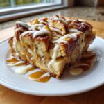 A close-up of a moist slice of apple bread pudding topped with caramel and vanilla icing.