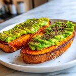 Close-up of two slices of vibrant orange sweet potato toast topped with fanned avocado slices and everything bagel seasoning.