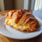 A golden brown baked ham and cheese croissant oozing melted cheese, sitting on a white plate.
