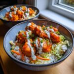 Close-up of a buffalo chicken bowl featuring crispy orange chicken pieces over rice and slaw, drizzled with ranch dressing.