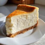A thick, creamy slice of baked cheesecake recipe on a white plate, showing a golden brown top and graham cracker crust.