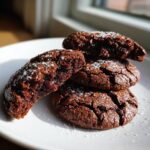 A stack of four rich, dark chocolate brownie cookies sprinkled with coarse sugar, showing a fudgy interior.
