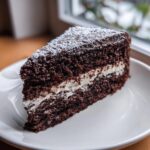 A triangular slice of dark chocolate yule log cake layered with white cream filling and dusted with powdered sugar.