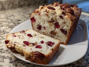 A close-up of a freshly baked cranberry walnut bread loaf, with one slice cut and resting beside it on a white platter.