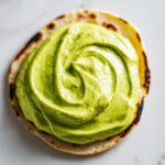 Close-up of vibrant green, creamy avocado sauce swirled on a small, charred tortilla.
