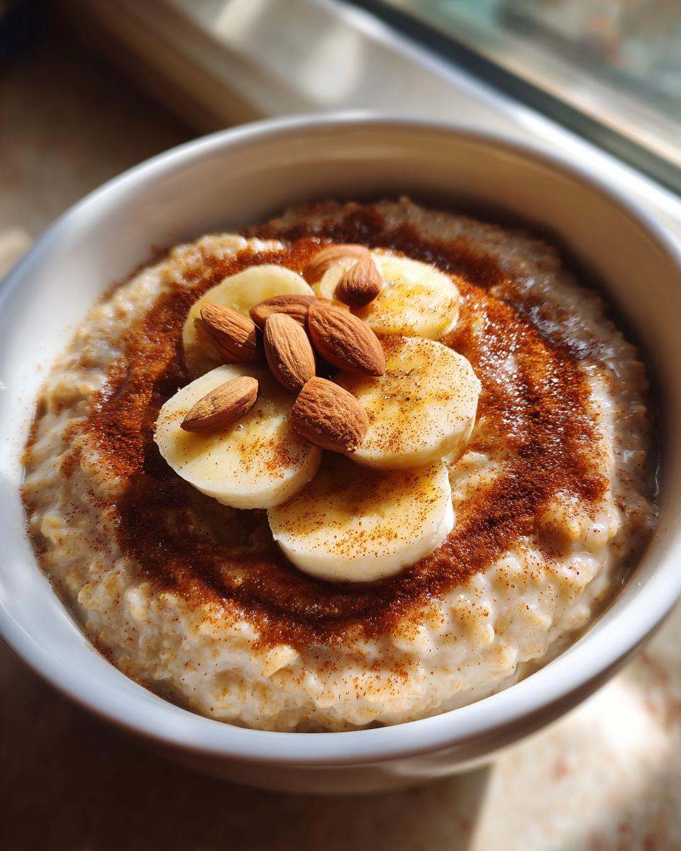 A close-up of creamy banana oatmeal topped with sliced bananas, whole almonds, and a dusting of cinnamon.