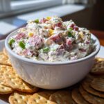 A white bowl filled with creamy hoagie dip, featuring chunks of meat and vegetables, surrounded by crackers.