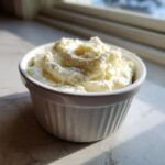 A small white ramekin filled with creamy, whipped horseradish sauce, sitting on a marbled surface near a window.