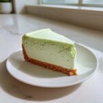 A perfect slice of light green key lime cheesecake with a thick graham cracker crust sits on a white plate.