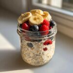A glass jar filled with creamy overnight oats, topped generously with fresh blueberries, raspberries, sliced banana, and sesame seeds.