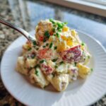 A spoonful of creamy red potato salad topped with fresh chives, lifted above a white plate.