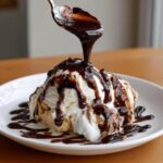 A spoonful of rich chocolate syrup being poured over scoops of vanilla ice cream served on a white plate.