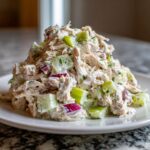 A mound of creamy dill pickle chicken salad featuring shredded chicken, diced celery, and red onion pieces on a white plate.