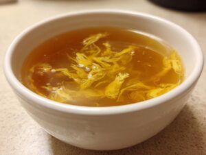 A close-up shot of a white bowl filled with hot, savory egg drop soup, showcasing the delicate yellow egg ribbons.