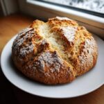 A golden-brown, rustic loaf of homemade soda bread with a cracked top dusted with flour, sitting on a white plate.