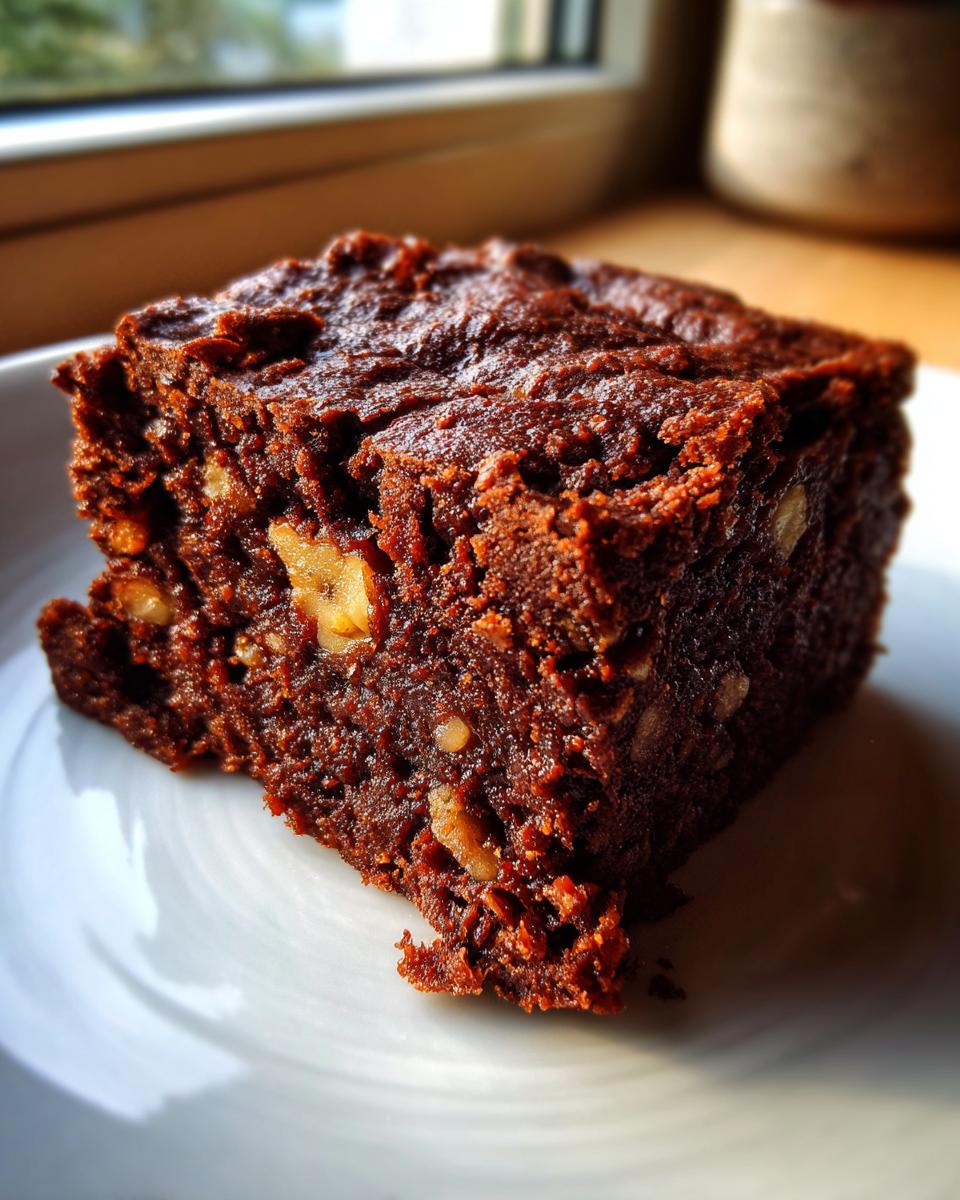 Close-up of a fudgy, dark brown date brownies square with visible chunks of walnuts, resting on a white plate.