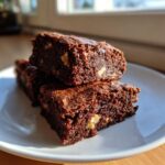 Close-up of stacked, fudgy date brownies studded with walnuts, sitting on a white plate in bright sunlight.