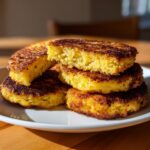 A stack of golden brown, perfectly pan-fried cornbread rounds, with one piece broken open to show the crumb.