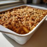 Close-up of a freshly baked apple crisp recipe with a golden, crunchy oat topping in a white rectangular baking dish.