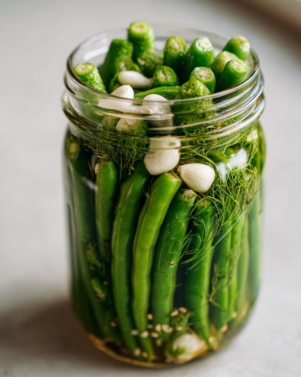 A glass jar packed tightly with fresh green dilly beans, whole garlic cloves, and dill fronds, ready for pickling.