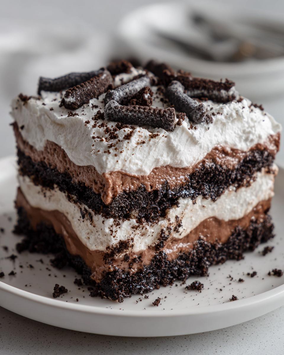 A square slice of layered dirt cake featuring dark chocolate cookie crust, chocolate pudding, whipped cream, and topped with crushed cookies.