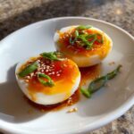 Two halves of soft-boiled marinated eggs drizzled with sauce, topped with sesame seeds and green onions.