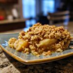 A close-up shot of a warm serving of apple crisp showing soft baked apples and a thick, golden brown oat crumble topping.