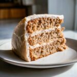 A close-up slice of moist, layered applesauce cake with vanilla bean frosting sitting on a light plate.